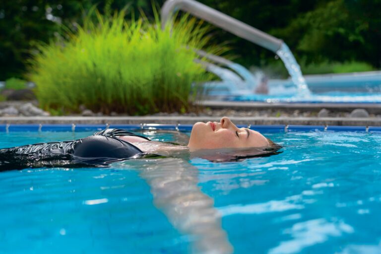 Eine Frau treibt entspannt im klaren Thermalwasser eines Außenpools, während im Hintergrund Wasser aus einer Edelstahl-Dusche fließt.